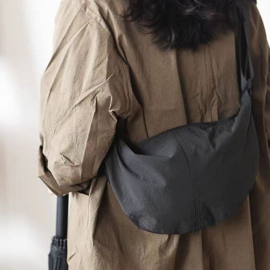A person wearing a brown dress is showcasing a black nylon crossbody bag with a wide shoulder strap. The person is holding a red smartphone inside the bag.